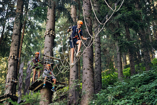 Teenage girl aged 16 with her brothers aged 13, wearing helmets and harnesses are walking in the outdoors ropes course adventure park. Kids are playing, laughing and having fun.\nCanon R5