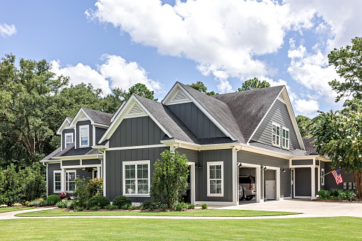 A large gray craftsman new construction house with a landscaped yard and leading pathway sidewalk on a sunny day with blue skies and clouds.