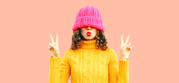 Winter colorful portrait of beautiful young woman covering her eyes with pink hat blowing her lips wearing yellow knitted sweater on background