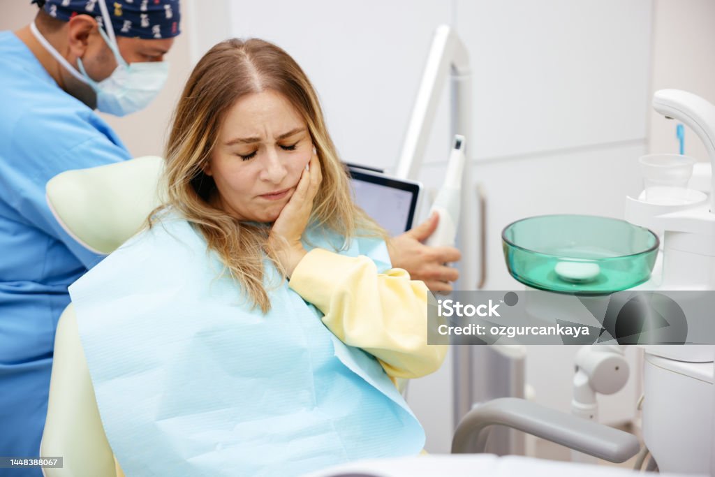 Young woman feeling pain, holding her cheek with hand at dentist's office.Toothache concept. Upset sad woman is holding hand on her cheek, teeth, sitting in chair in dental office, clinic. Female is feeling pain and toothache, waiting for doctor dentist examining. Dentistry concept. Accidents and Disasters Stock Photo