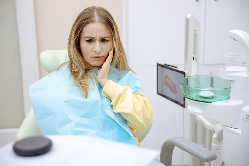 Upset sad woman is holding hand on her cheek, teeth, sitting in chair in dental office, clinic. Female is feeling pain and toothache, waiting for doctor dentist examining. Dentistry concept.