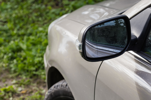 Side view mirror of the gray car with integrated turn signal repeater standing on the meadow, rear view on a blurred background of car front part, close-up in selective focus