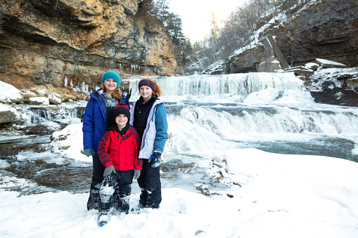 Two sisters and their younger brother smiling at the camera on a winter day. Waterfalls can be seen behind them, falling into the river around them.