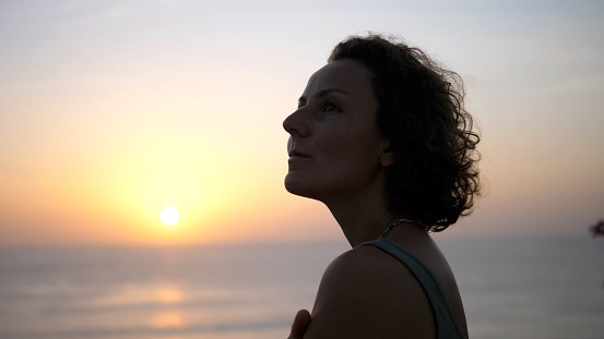 In the background, the sun hides behind the sea horizon. The last rays of the orange-yellow sun illuminate the silhouette of a woman on the ocean. The woman hugs her bare shoulders.