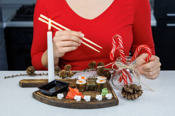 Japanese rolls with red fish on a wooden cutting board. A girl in a red sweater eats sushi with chopsticks. Table decoration with candles and sweets. Christmas concept Japanese rolls with red fish on a wooden cutting board. A girl in a red sweater eats sushi with chopsticks. Table decoration with candles and sweets. Christmas concept. kids eating tuna stock pictures, royalty-free photos & images