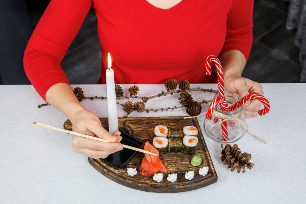 Japanese rolls with red fish on a wooden cutting board. A girl in a red sweater eats sushi with chopsticks. Table decoration with candles and sweets. Christmas concept Japanese rolls with red fish on a wooden cutting board. A girl in a red sweater eats sushi with chopsticks. Table decoration with candles and sweets. Christmas concept. kids eating tuna stock pictures, royalty-free photos & images