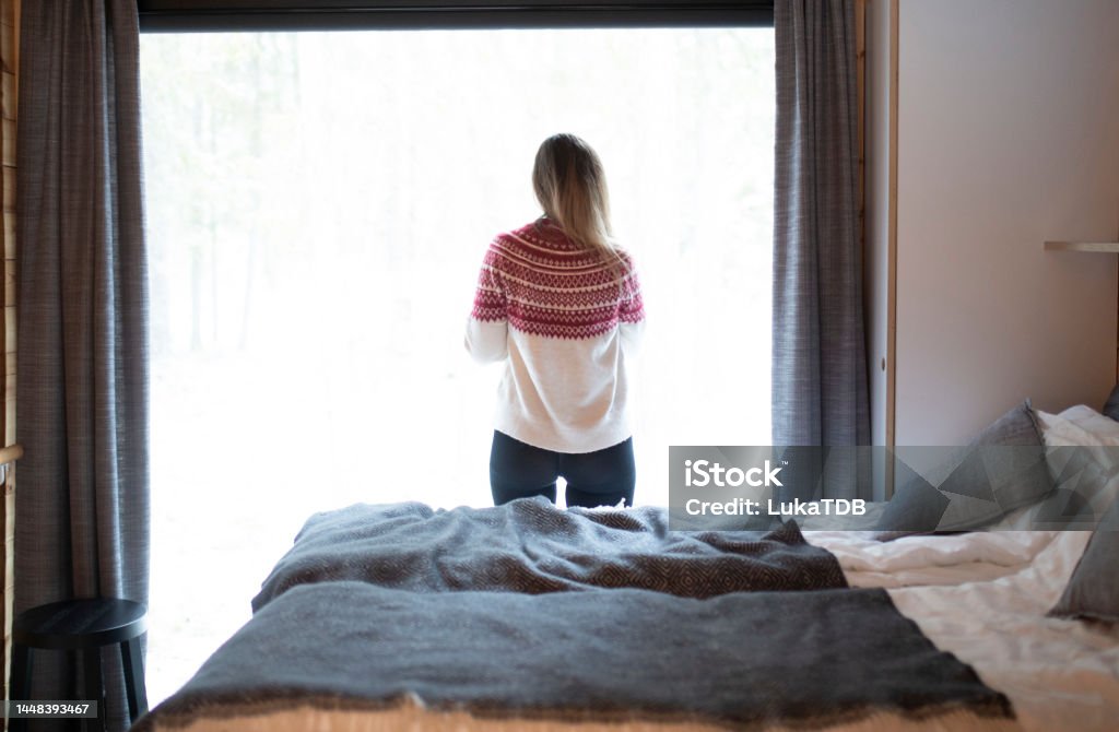 An unrecognizable woman in winter sweater looks out the window Rear view of a woman standing in a cottage looking out the window Cottage Stock Photo