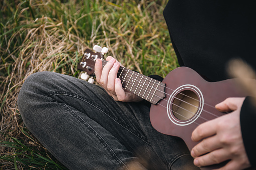 A man plays the ukulele guitar in nature, close-up fingers pinch the strings, blurred background, copy space.