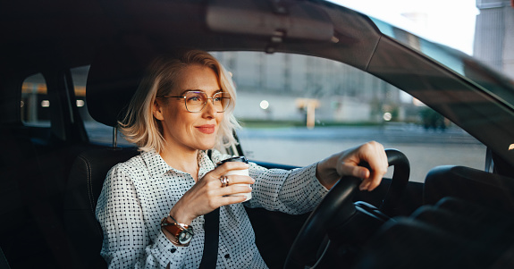 Smiling business woman holding disposable cup with hot drink while traveling with her car around the city.