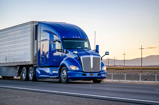 Industrial comfort big rig powerful blue semi truck tractor with high cab transporting frozen commercial cargo in refrigerator semi trailer running on the highway road at sunset in California