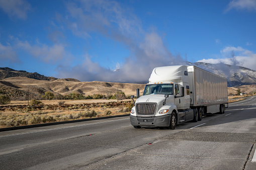 Industrial long hauler big rig white semi truck tractor with chrome parts transporting commercial cargo in loaded dry van semi trailer running on the highway road at sunny day in California