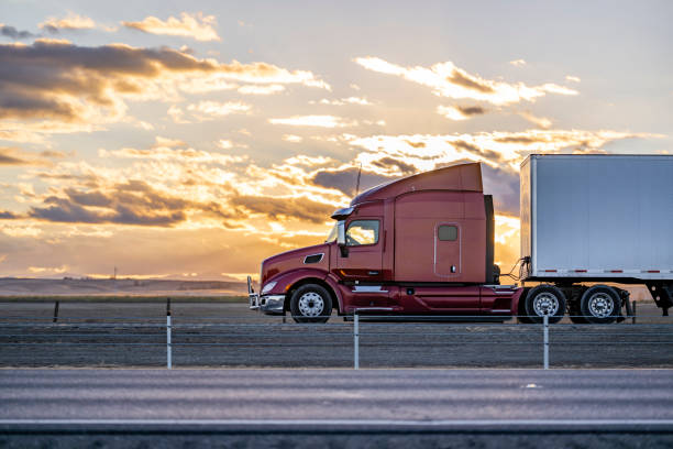 Burgundy big rig semi truck with grille guard and high cab spoiler transporting cargo in dry van semi trailer driving on the road at twilight stock photo