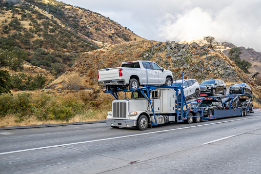 Industrial big rig white car hauler semi truck transporting cars on special two level hydraulic modular semi trailer driving on the highway road with snow mountains hidden in the clouds in California
