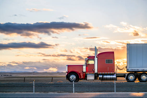 Side view of red big rig classic semi truck transporting cargo in dry van semi trailer moving on the road at sunset stock photo