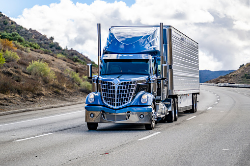 Industrial grade popular big rig blue semi truck tractor with chrome parts transporting frozen commercial cargo in refrigerator semi trailer running on the highway road at sunny day in California