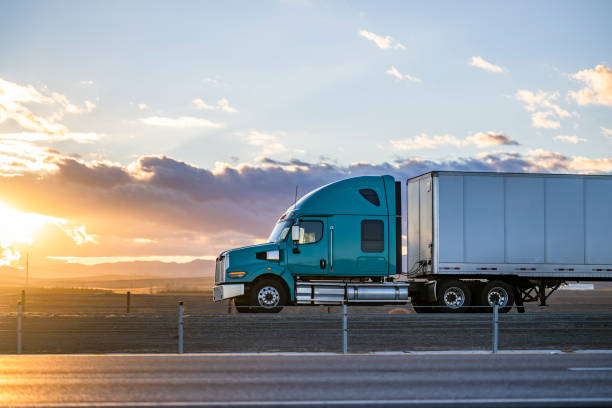 Streamline green big rig bonnet semi truck transporting cargo in dry van semi trailer driving on the highway road with fields on the sides at sunset time stock photo
