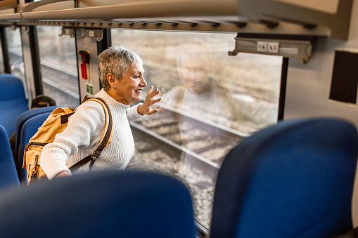 Mature woman with short grey hair and backpack traveling by train, standing and lookingthrough the window, impatiently waiting for next stop.