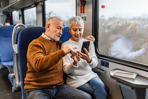 Mature heterosexual couple traveling by train, sitting next to each other, using smart phone, taking a selfie, enjoying ride.