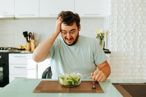 Holding his head, a young man looks at his salad showing the grueling pangs of the diet. The guy can no longer see the greens and sit on a plant based diet. Stop diet.