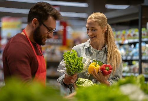Sellers talk to smiling customers and help them choose food in a large store with fresh vegetables. Mixed races between buyers and sellers.