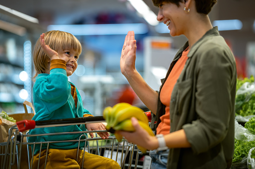 A smiling mother with a happy little child sitting in a shopping cart chooses vegetables and fruits. The child actively helps her. His mother teaches him that vegetables and fruits are good for him.