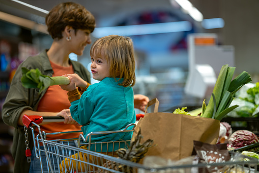 A smiling mother with a happy little child sitting in a shopping cart chooses vegetables and fruits. The child actively helps her. His mother teaches him that vegetables and fruits are good for him.