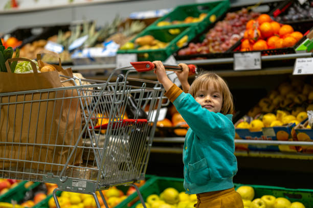 A smiling mother with a happy little child sitting in a shopping cart chooses vegetables and fruits. The child actively helps her. His mother teaches him that vegetables and fruits are good for him.