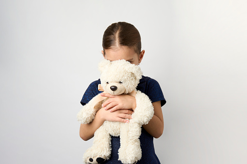 A little curly haired girl sits up on an exam table, as she hugs her teddy bear tightly for comfort.  She is dressed casually in a pink striped shirt and smiling gently as she waits for the doctor to enter the room.