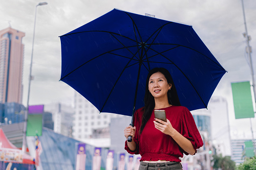 Beautiful woman holding blue umbrella in rainy day holding smartphone in city