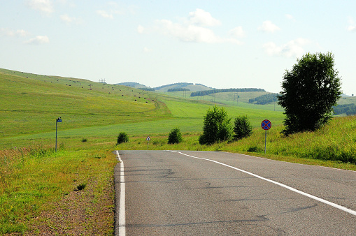 A two-lane asphalt road curves through a picturesque hilly valley on a warm summer day. Khakassia, Siberia, Russia.