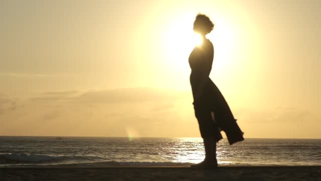 Time lapse of a silhouette of a woman in a midi dress, standing sideways to the camera against the backdrop of a sunset and seashore. The sun creates a rainbow aura around the woman.