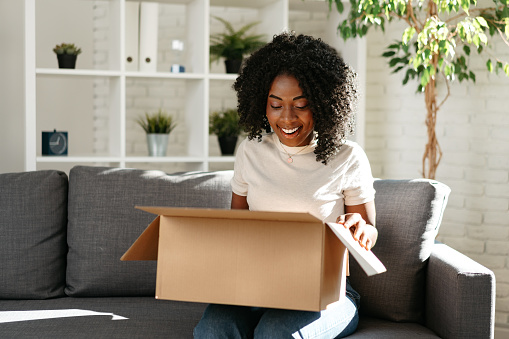 Young african woman sit on couch at home unpacking parcel cardboard box with online purchase, close up