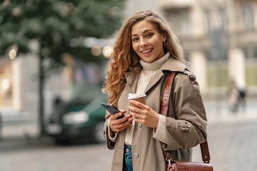 Young woman with curly blonde hair using the phone with a cup of coffee in hands on the city streets, portrait