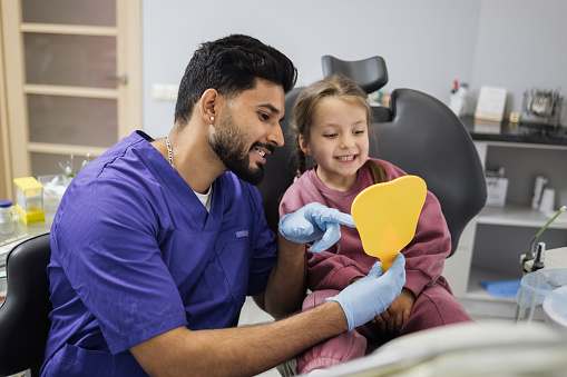 Happy cute little girl enjoying her beautiful toothy smile looking at mirror while young bearded confident male dentist holding it in modern light dental office clinic.