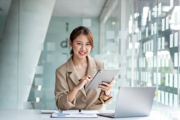 Portrait of smiling young beautiful Asian businesswoman sitting with laptop computer looking at camera in the office. stock photo