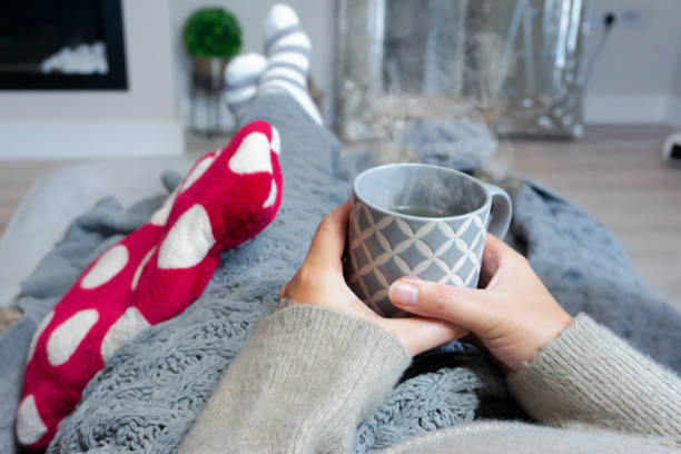 Laying on a sofa while having a hot drink and staying warm Staying warm with a hot water bottle, blanket and a hot drink, taken in the UK 2022 hibernation stock pictures, royalty-free photos & images