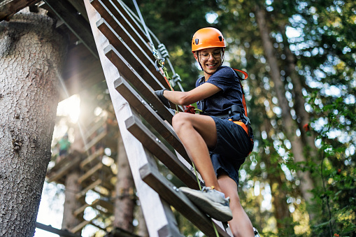Teenage boy aged 13, wearing helmet and harness is climbing up the ladder in the outdoors ropes course adventure park. The boy is looking over shoulder and laughing.\nCanon R5