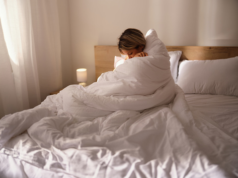 Young sad woman feeling lonely while being covered with duvet on a bed in bedroom. Photographed in medium format.