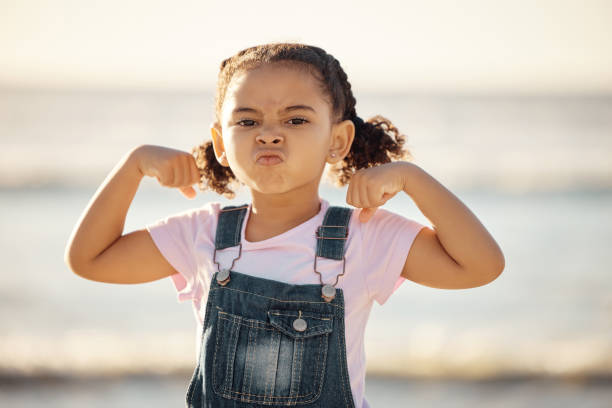 beach, summer and little girl pulling funny face and flexing on holiday in mexico. strong, courage and child at ocean on vacation in cancun. waves, sea and sand, healthy kid in nature on school break - flexing muscles fotos imagens e fotografias de stock