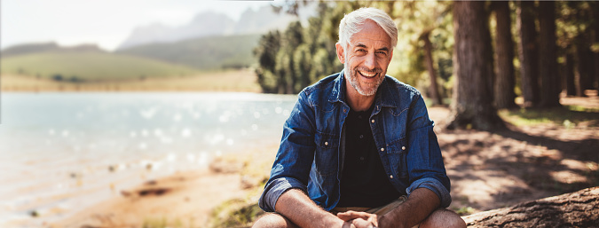 Portrait of a happy mature man sitting near a lake looking at camera and smiling. The senior caucasian man is sitting on a log by the lake on a summer day. Concept of healthy senior lifestyle and outdoor camping and living.