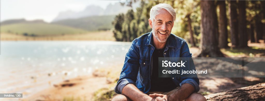 Happy and healthy senior man smiling while enjoying an active lifestyle in nature and outdoor camping Portrait of a happy mature man sitting near a lake looking at camera and smiling. The senior caucasian man is sitting on a log by the lake on a summer day. Concept of healthy senior lifestyle and outdoor camping and living. Mature Men Stock Photo