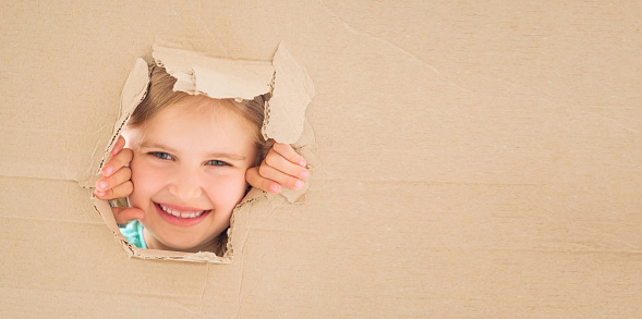 Playful little girl gazing from cardboard box hole right in front of her.