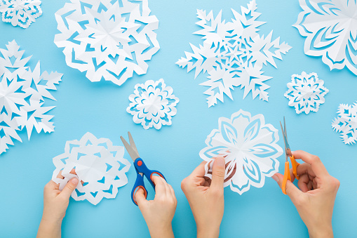 Adult mother and child hands cutting different white snowflake shapes from paper on light blue table background. Pastel color. Making decoration elements for winter festive. Closeup. Top down view.