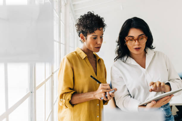 two business women having a discussion, they're standing in an office and using a tablet - meeting document working people imagens e fotografias de stock