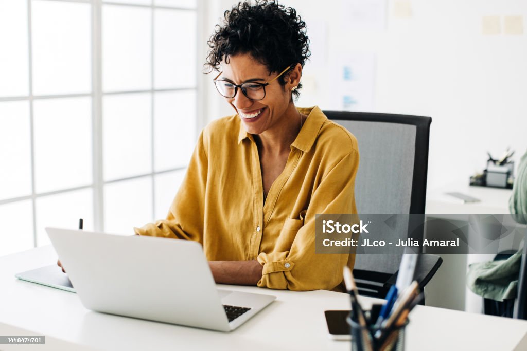 Graphic designer smiles as she works on a laptop in an office Graphic designer smiles as she works on a laptop in an office. Woman using a graphics tablet to make drawing designs. Creative business woman enjoys working on her project in an office. Women Stock Photo