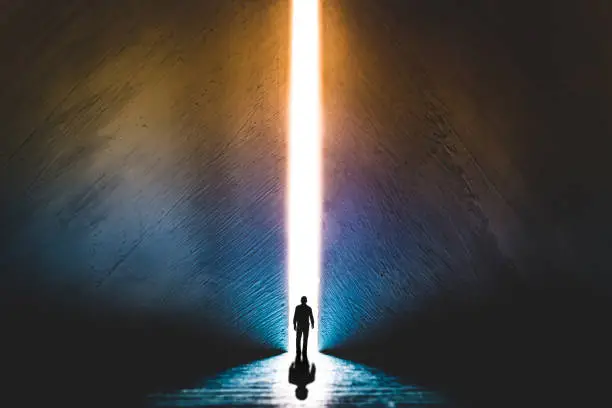 Photo of The silhouette of a man walking towards a bright light in the opened huge wall. A light in the end of a tunnel. The concept of success, freedom of choice, open mind, meditation.