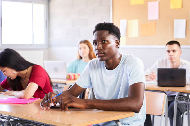 African American student attending teacher lesson at classroom. Black guy in class study with a multiracial high school college. Young boys and girls sitting at desk. Education concept stock photo