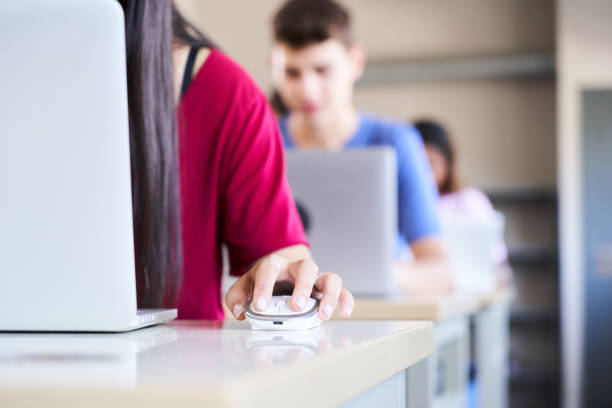 Close-up of a hand clutching a mouse in a high school classroom, a group of cheerful students study with a laptop computer. Concept of technology and education stock photo