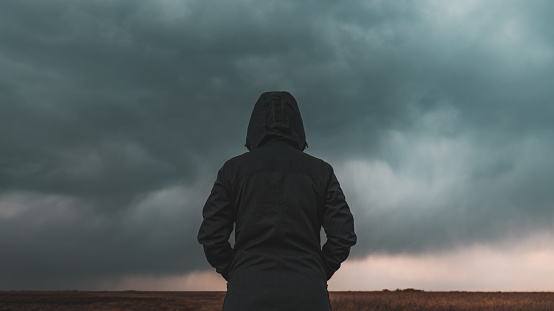 Rear view of female person wearing hooded jacket against dark moody dramatic clouds at sky, woman looking into uncertain ominous future