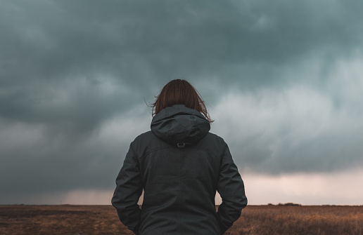 Woman standing in meadow, looking at the horizon and dark dramatic stormy clouds, rear view selective focus